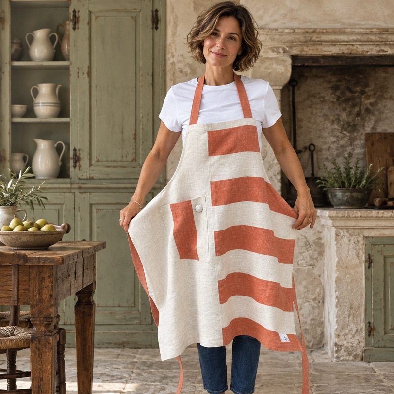 Terracotta Striped Linen Apron – Follia Pura-La-Bottega-di-Casa Model holds out the sides of the Follia Pura ecru linen apron with terracotta stripes and a contrasting pocket in a rustic kitchen.