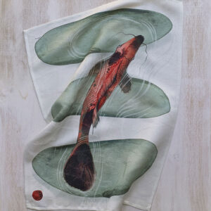 Linen and cotton kitchen towel with red koi carp ink wash illustration laid on a light wood surface — single red carp among smooth grey stones. L'Arcolaio for La Bottega di Casa.