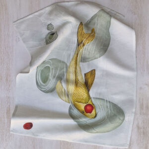 Linen and cotton kitchen towel with golden koi carp ink wash illustration laid on a light wood surface — large golden carp with red mark among smooth grey stones.