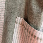 Close-up of the natural green linen fabrics used in the Follia Pura apron, showing herringbone and striped weaves.